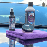 Three car care products from Squid Brand are displayed on a purple microfiber cloth on a glossy blue car hood. The products include a bottle of Ultra Violet Ceramic Protect spray, a large bottle of HO Pure Vehicle Shampoo, and a tin of Asparagus Car Wax.